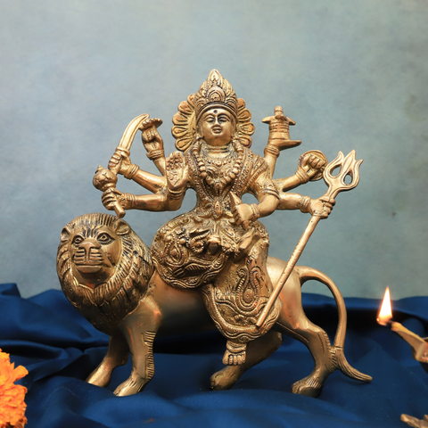 Front view of handcrafted brass Durga Murti with eight hands holding weapons, seated on a lion, displayed on a blue cloth with a lit diya.