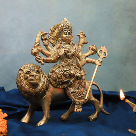 Handcrafted brass Durga Murti with eight hands riding a lion, holding traditional weapons and symbols, displayed on a blue cloth with a lit diya nearby