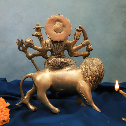 Back view of handcrafted brass Durga Murti with eight hands holding weapons, seated on a lion, displayed on a gold cloth with a lit diya.