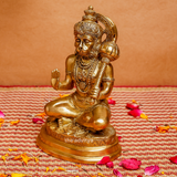 Side angle of a brass Hanuman idol in sitting posture holding a Gada, placed on a festive mat with flower petals.