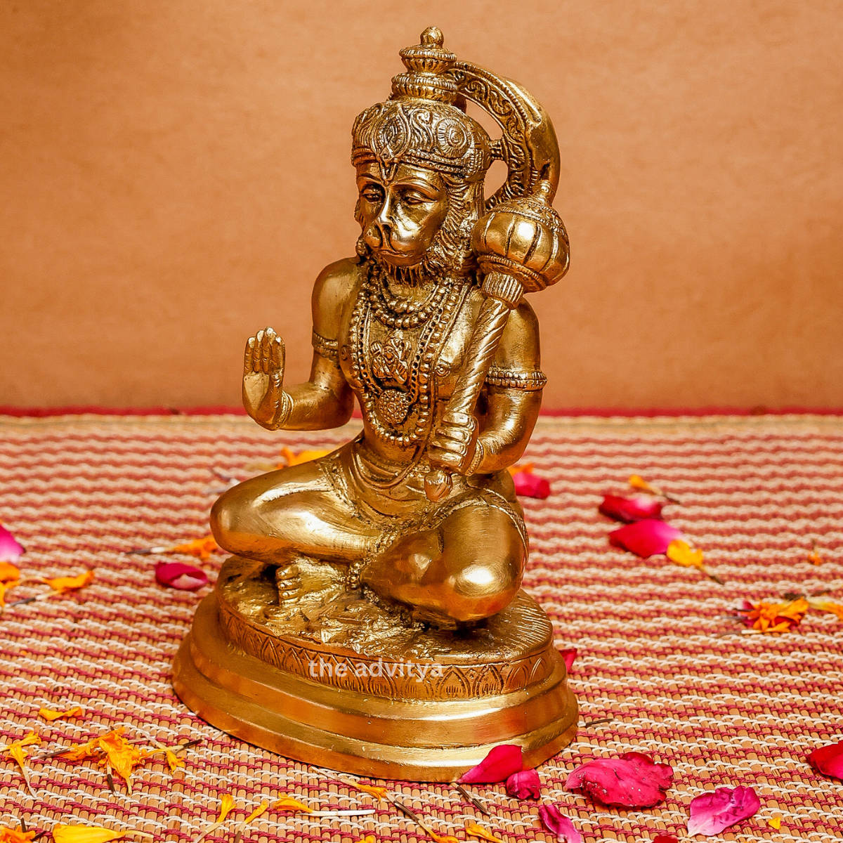 Side angle of a brass Hanuman idol in sitting posture holding a Gada, placed on a festive mat with flower petals.